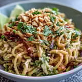 A vibrant Asian Peanut Noodle Bowl topped with chopped peanuts and sesame seeds, served with lime wedges on the side.