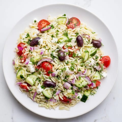 A close-up of Greek Orzo Salad in a white bowl, featuring al dente pasta, diced cucumber, halved cherry tomatoes, and crumbled feta cheese tossed in a bright lemon-oregano dressing.