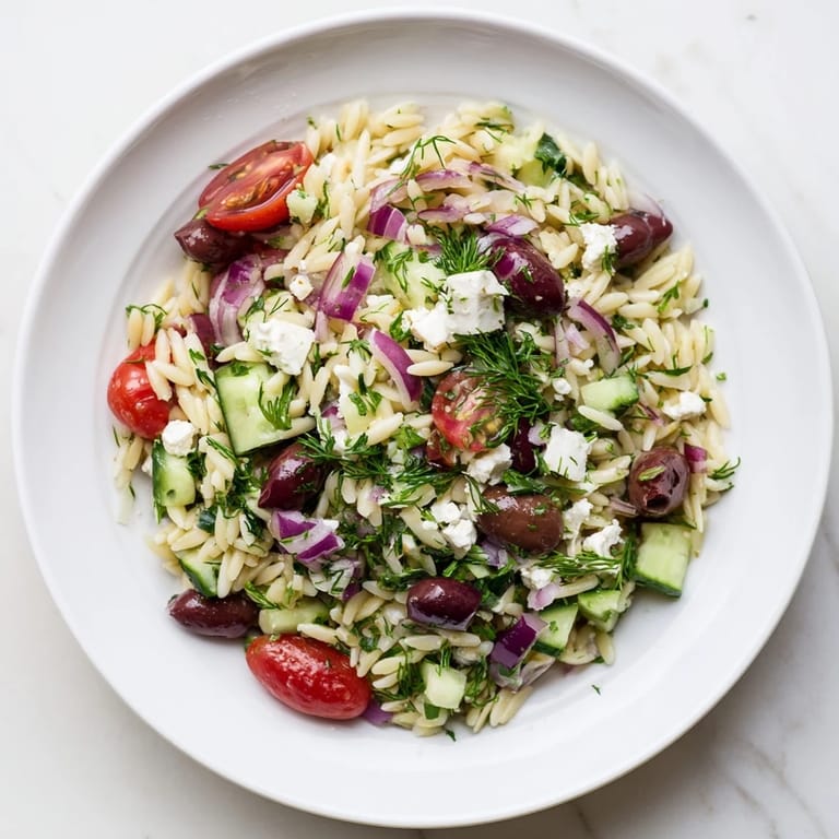A serving spoon lifts a portion of Greek Orzo Salad, revealing juicy tomatoes, crisp vegetables, and briny Kalamata olives mixed with tender pasta and fresh herbs.